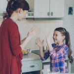 Happy young mother and cute funny daughter playing clapping hands game and have fun while cooking in kitchen at home. Family, cooking and people concept