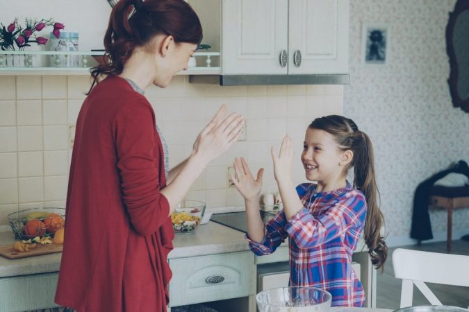 Happy young mother and cute funny daughter playing clapping hands game and have fun while cooking in kitchen at home. Family, cooking and people concept
