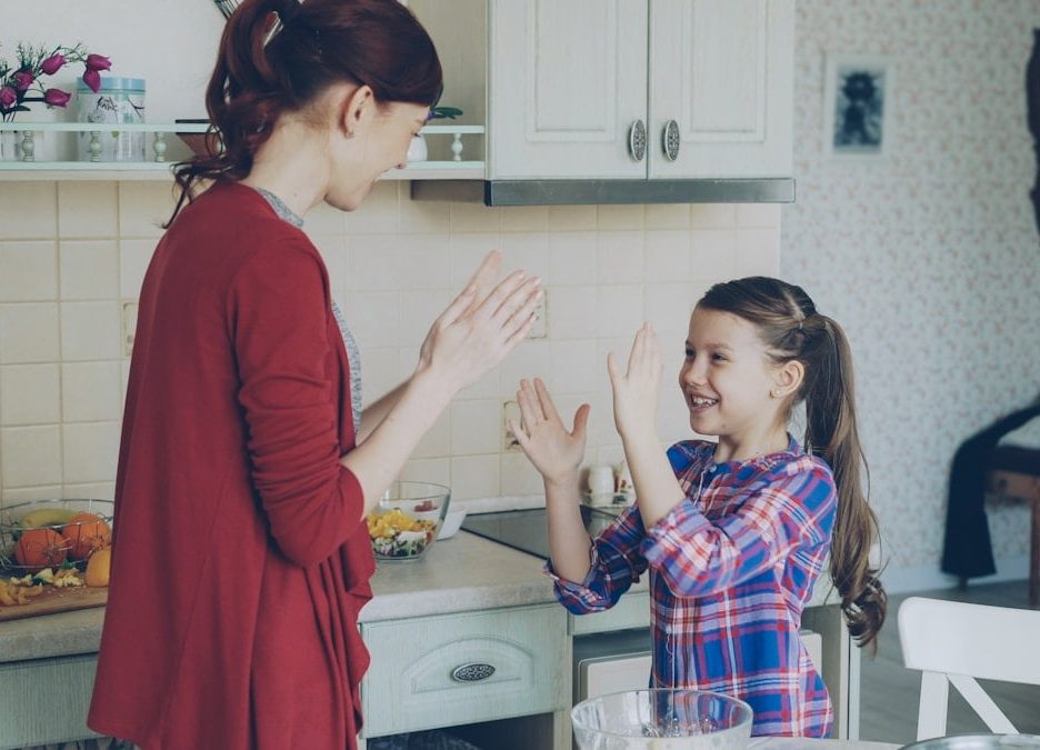 Happy young mother and cute funny daughter playing clapping hands game and have fun while cooking in kitchen at home. Family, cooking and people concept