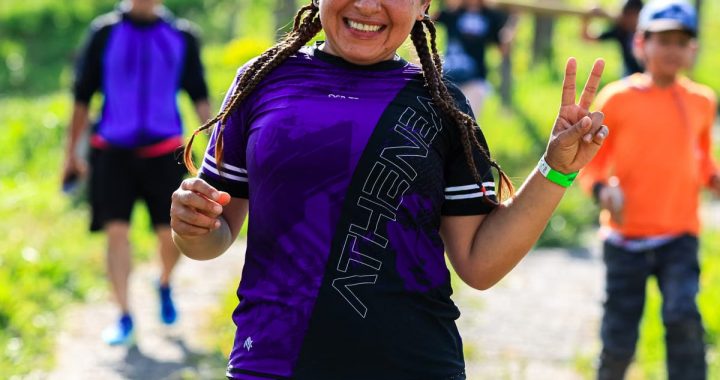 A cheerful female runner flashes a peace sign during an outdoor run with others in a sunny, green setting.