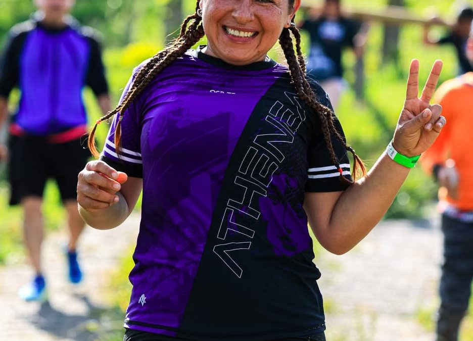 A cheerful female runner flashes a peace sign during an outdoor run with others in a sunny, green setting.